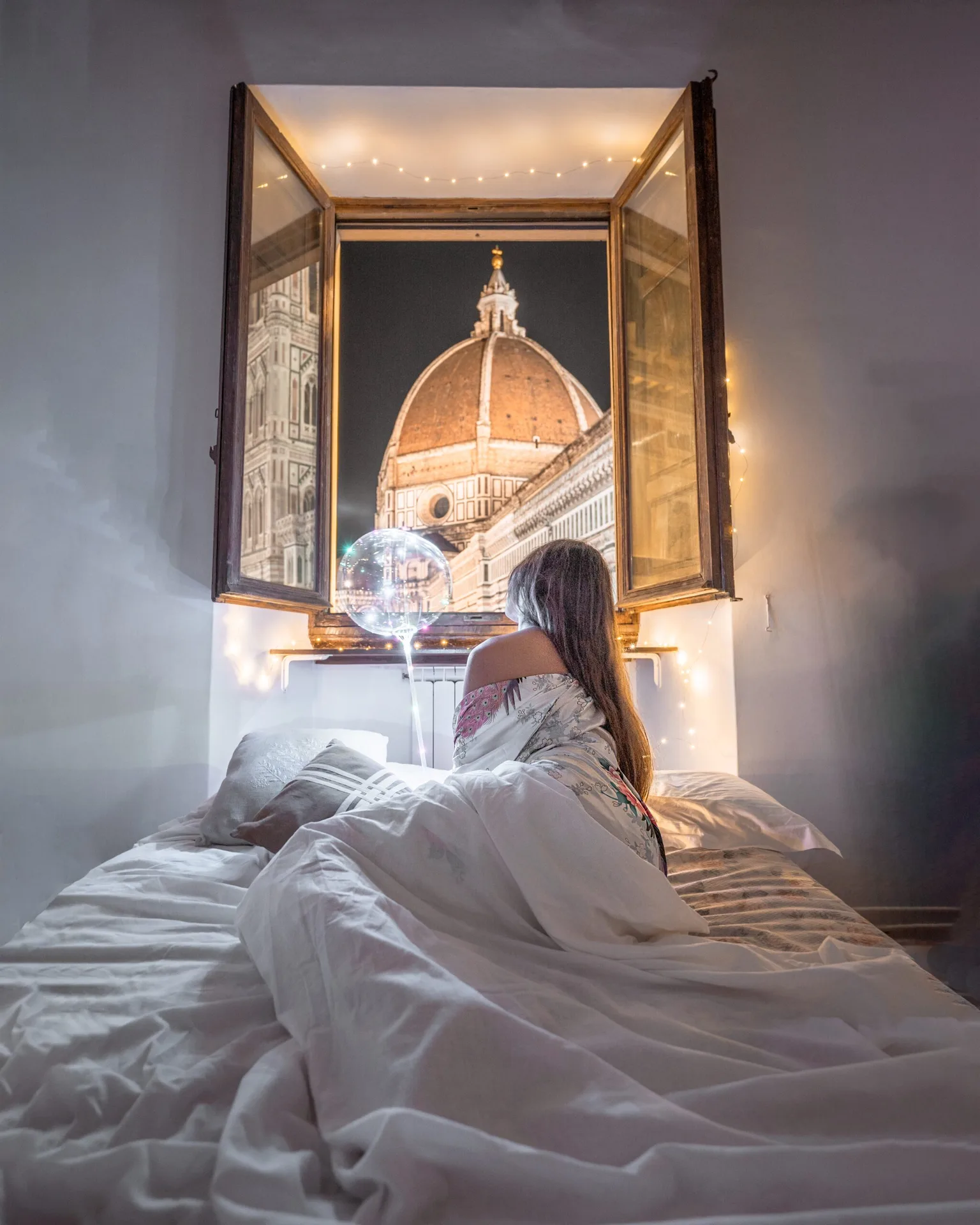 Woman wrapped in a blanket sits on a bed at an open window looking at Florence's illuminated Duomo dome at night.