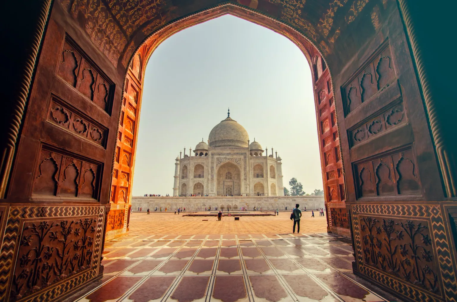 View of the Taj Mahal framed by an ornate red sandstone gateway with a tiled forecourt and visitors.