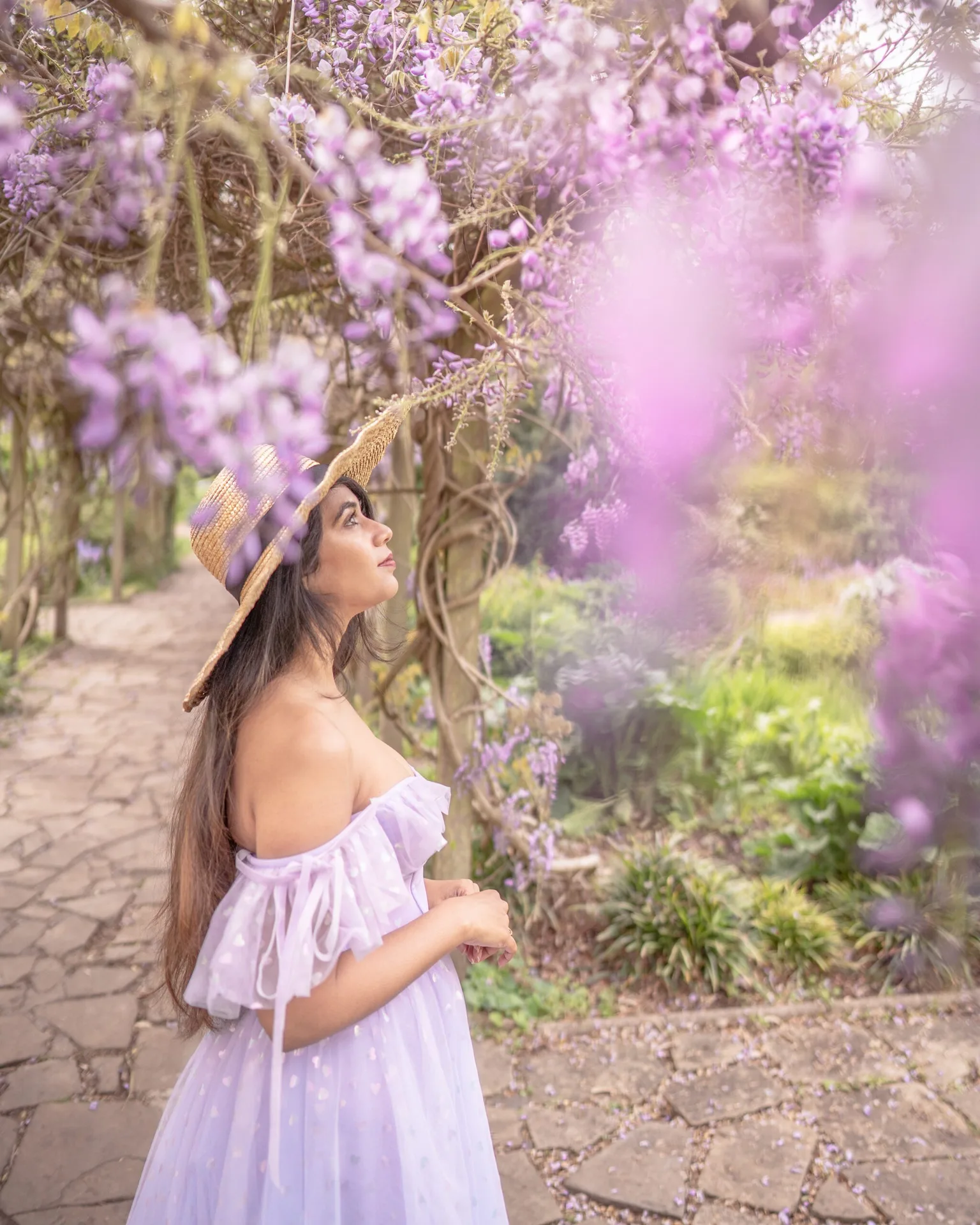 Woman in a lavender off-shoulder dress and straw hat gazing up at hanging purple wisteria along a garden path.