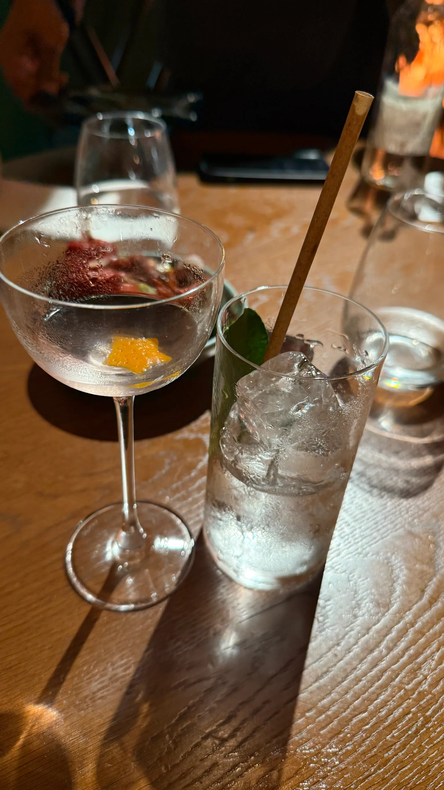 A cocktail glass with a citrus peel beside a tall iced glass with a lime leaf and bamboo straw on a wooden table.