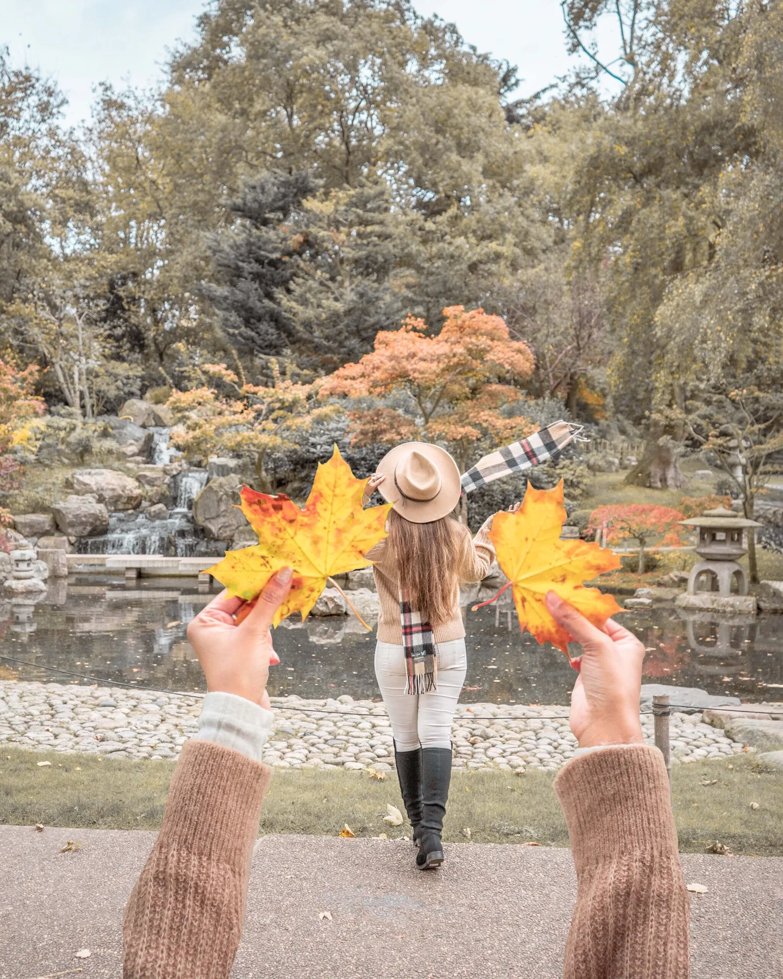 Woman in a hat and scarf walking toward a pond and waterfall framed by two large golden autumn leaves held in the foreground.