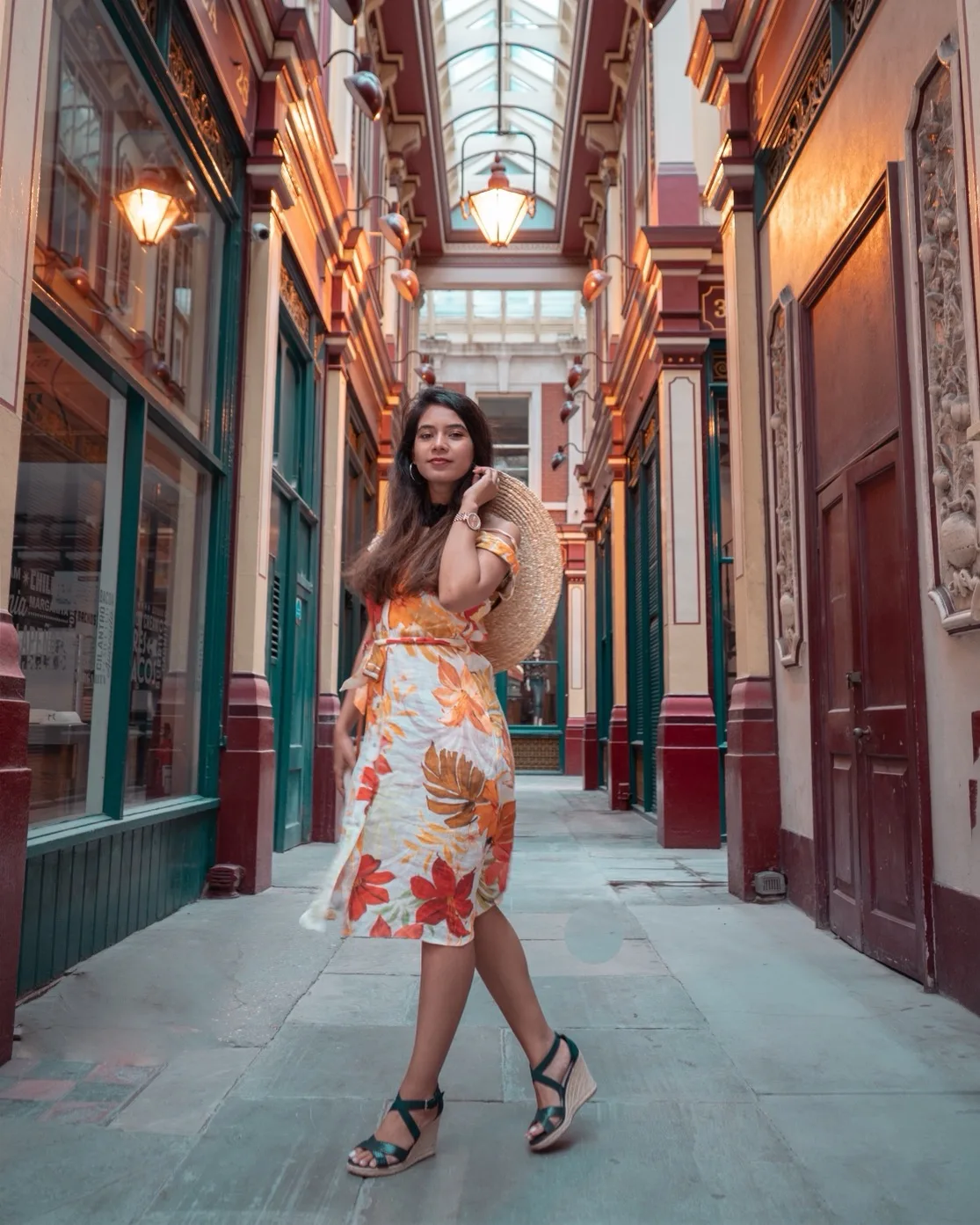 Woman in a floral summer dress and straw hat posing in a sunlit, ornate covered arcade with warm lamps and decorative columns