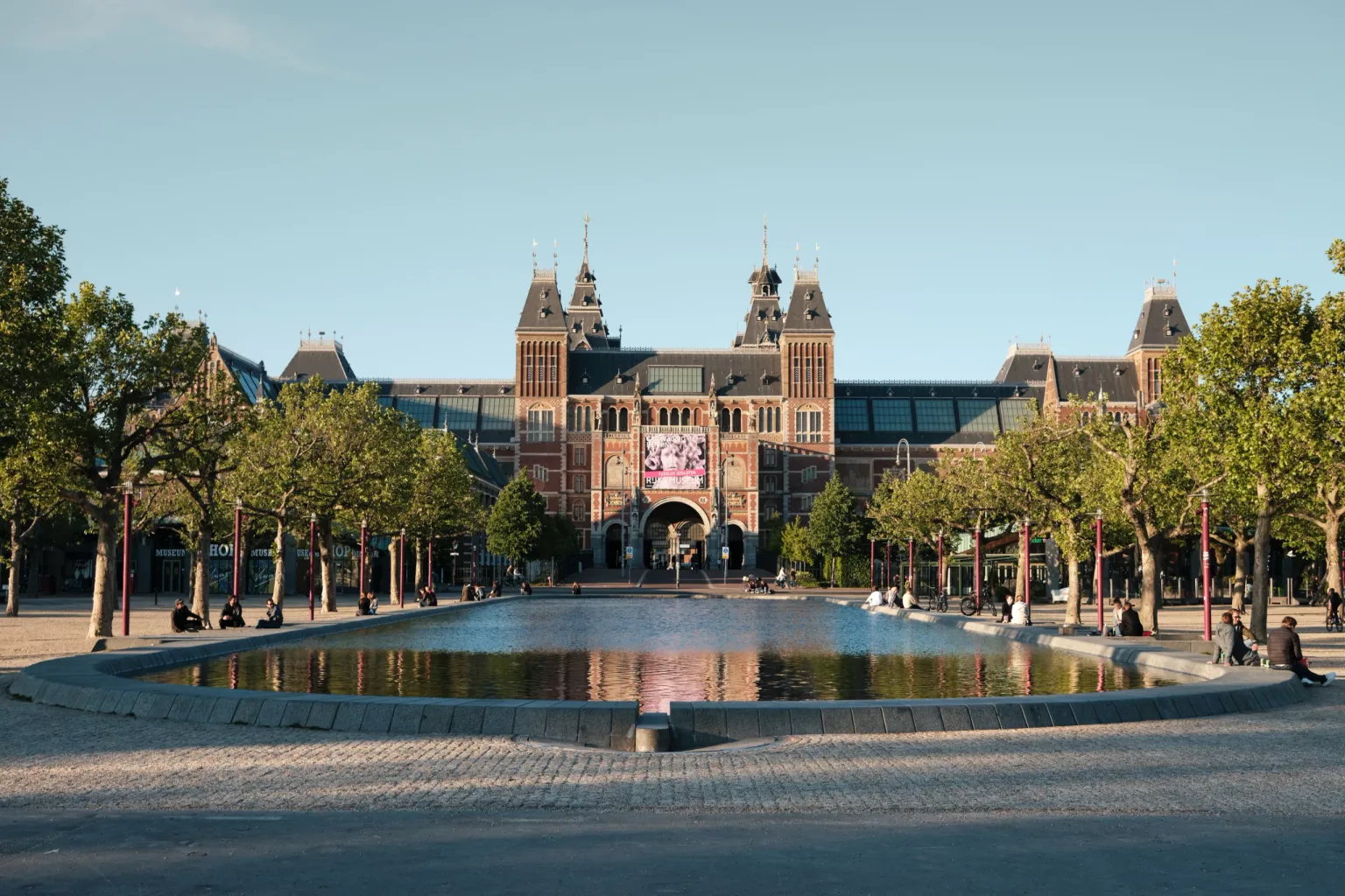 Grand museum building with ornate towers reflected in a long rectangular pond framed by trees and seating.