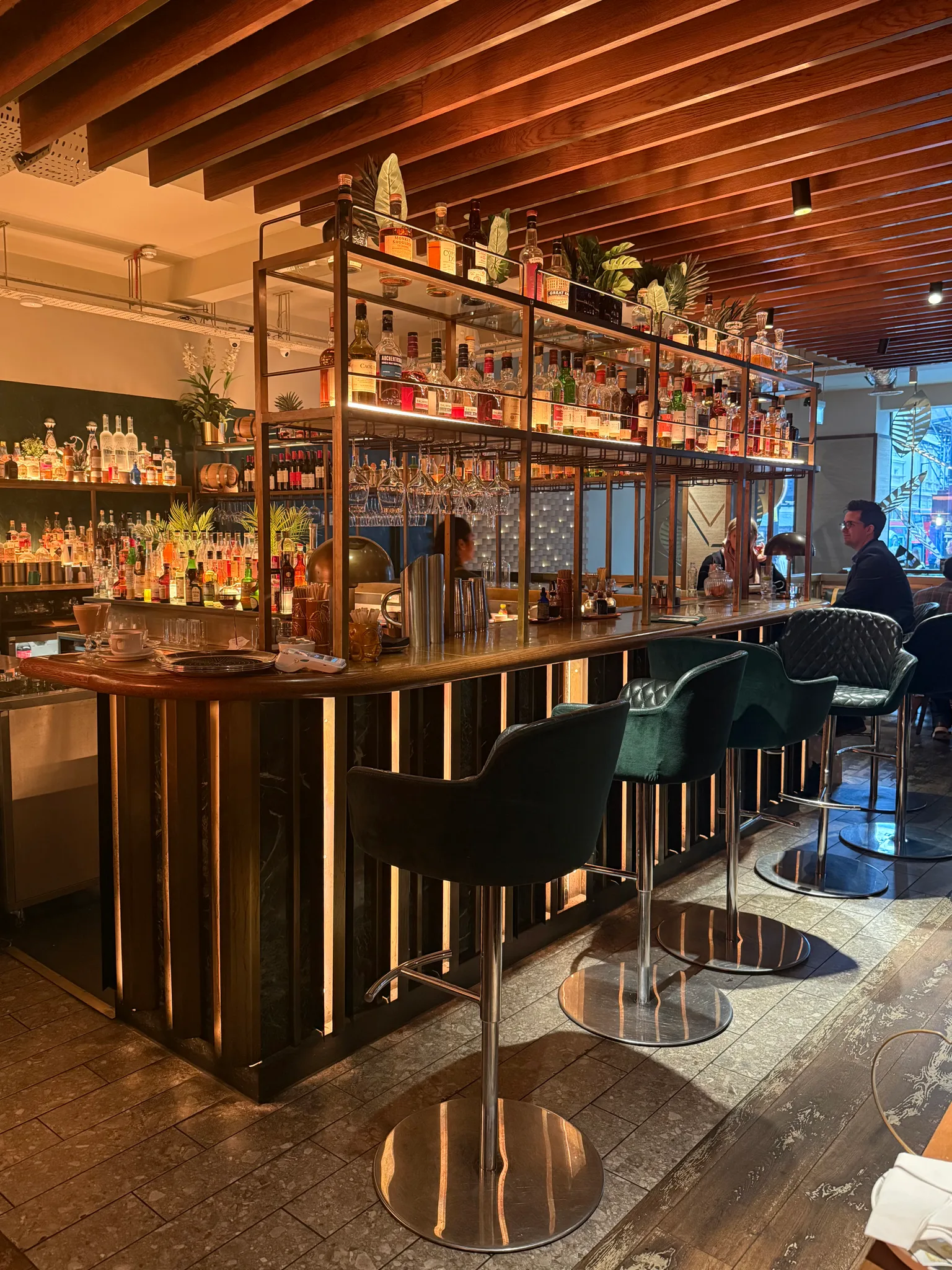 Stylish dimly lit hotel bar with illuminated shelving stocked with bottles and velvet bar stools along a wooden counter.