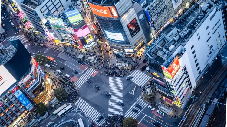 Aerial view of a busy city intersection surrounded by bright neon billboards and crowds at dusk