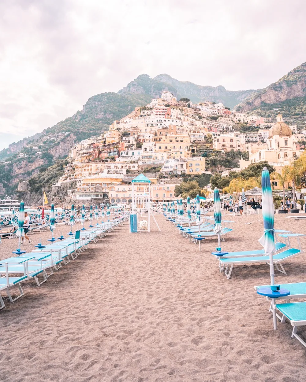 Sun-soaked Positano beach with rows of blue sunbeds and umbrellas in front of colourful cliffside houses.