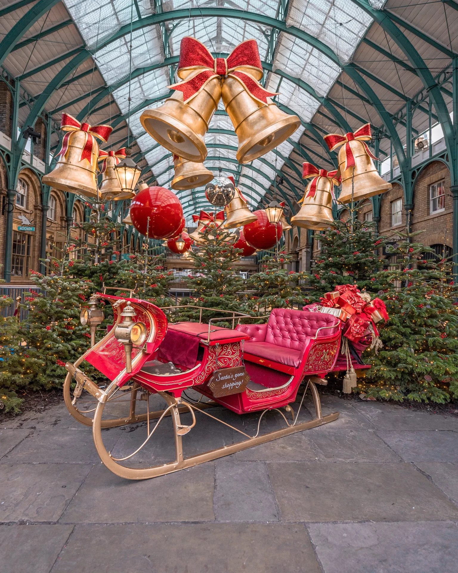 Decorative red sleigh surrounded by Christmas trees and giant golden bells hanging from a glass-roofed arcade.