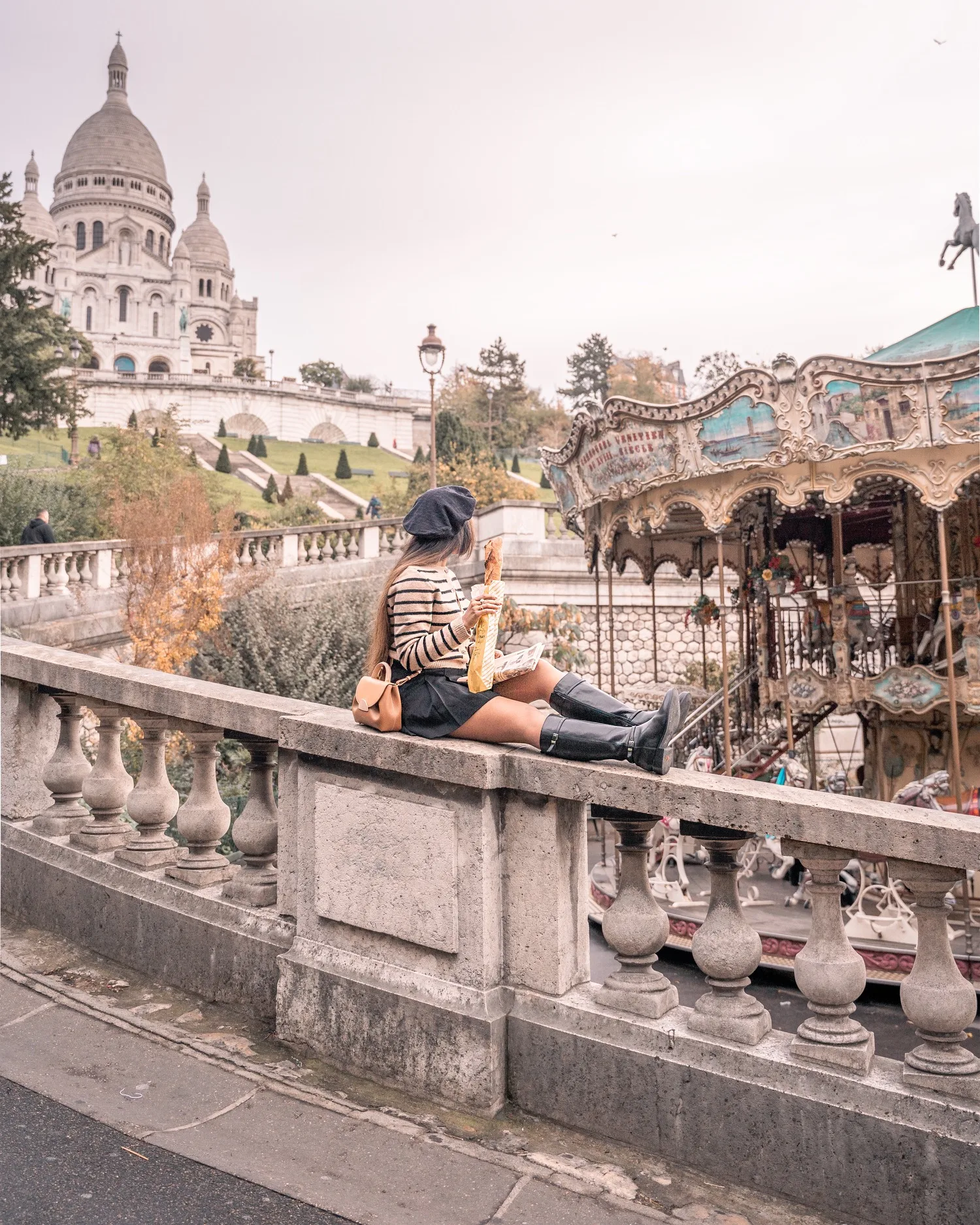 Woman in a beret eating a baguette on a stone balustrade overlooking Sacré‑Cœur and a vintage carousel in Paris.