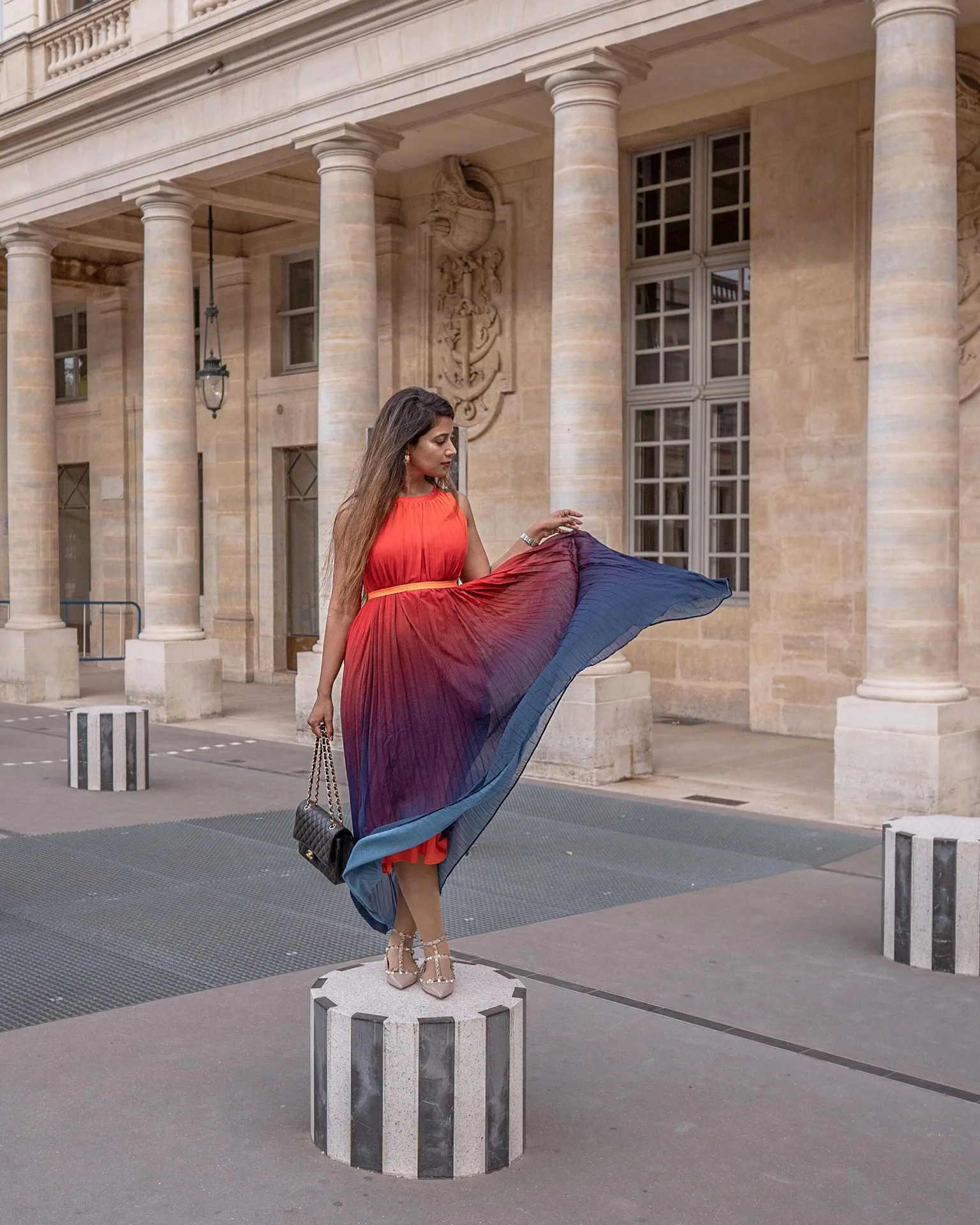 Woman in a flowing red-to-blue ombré dress poses on a striped stone column outside a classical columned building, holding a chain-strap bag.