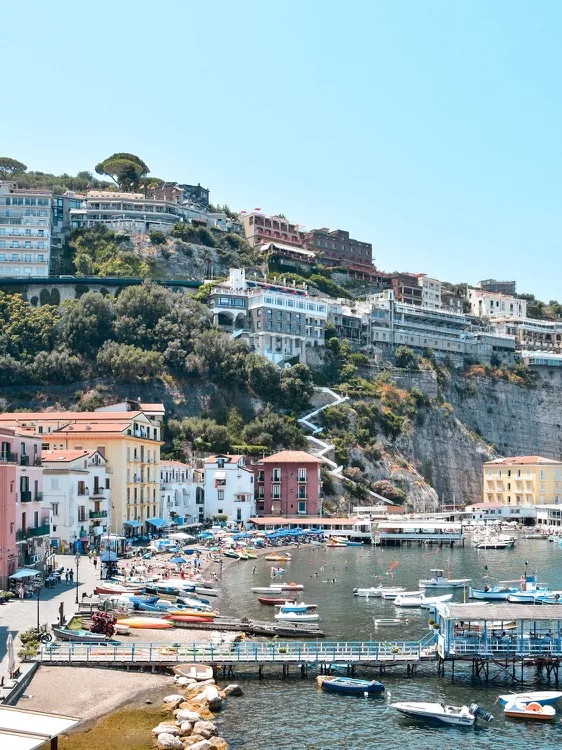 Colourful seaside town with boats in a protected harbour beneath cliffside villas and winding stairways.