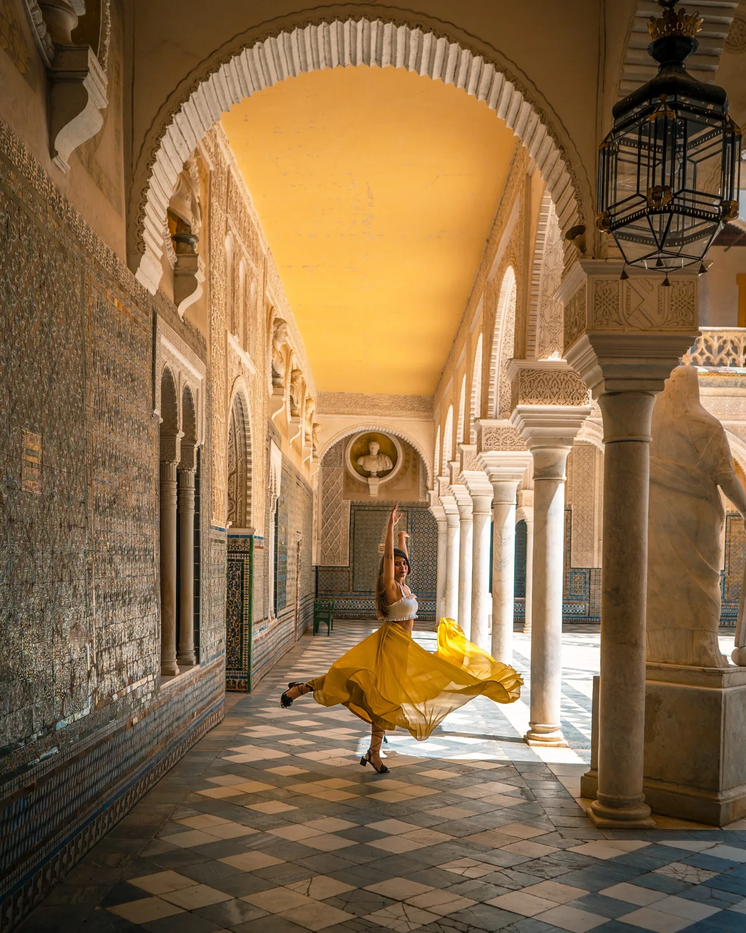 Woman in a flowing yellow skirt twirling in a sunlit, columned Moorish-style cloister with patterned tile floors.