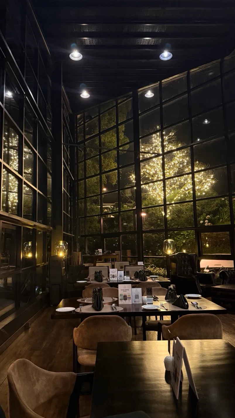 Cozy dimly lit restaurant with floor-to-ceiling glass walls looking out onto a tree wrapped in warm fairy lights.