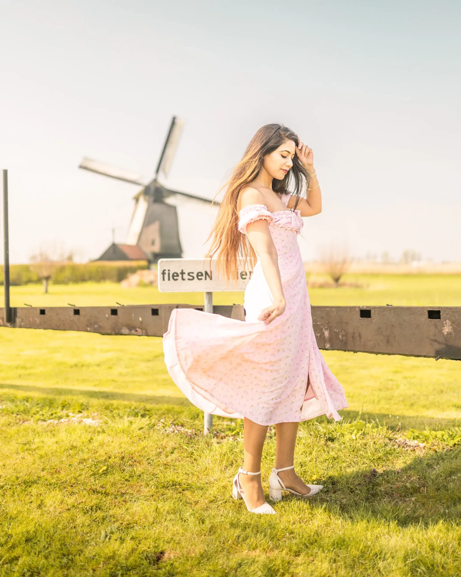 Young woman in a flowing pink dress twirling on green grass with a traditional Dutch windmill in the sunlit background.