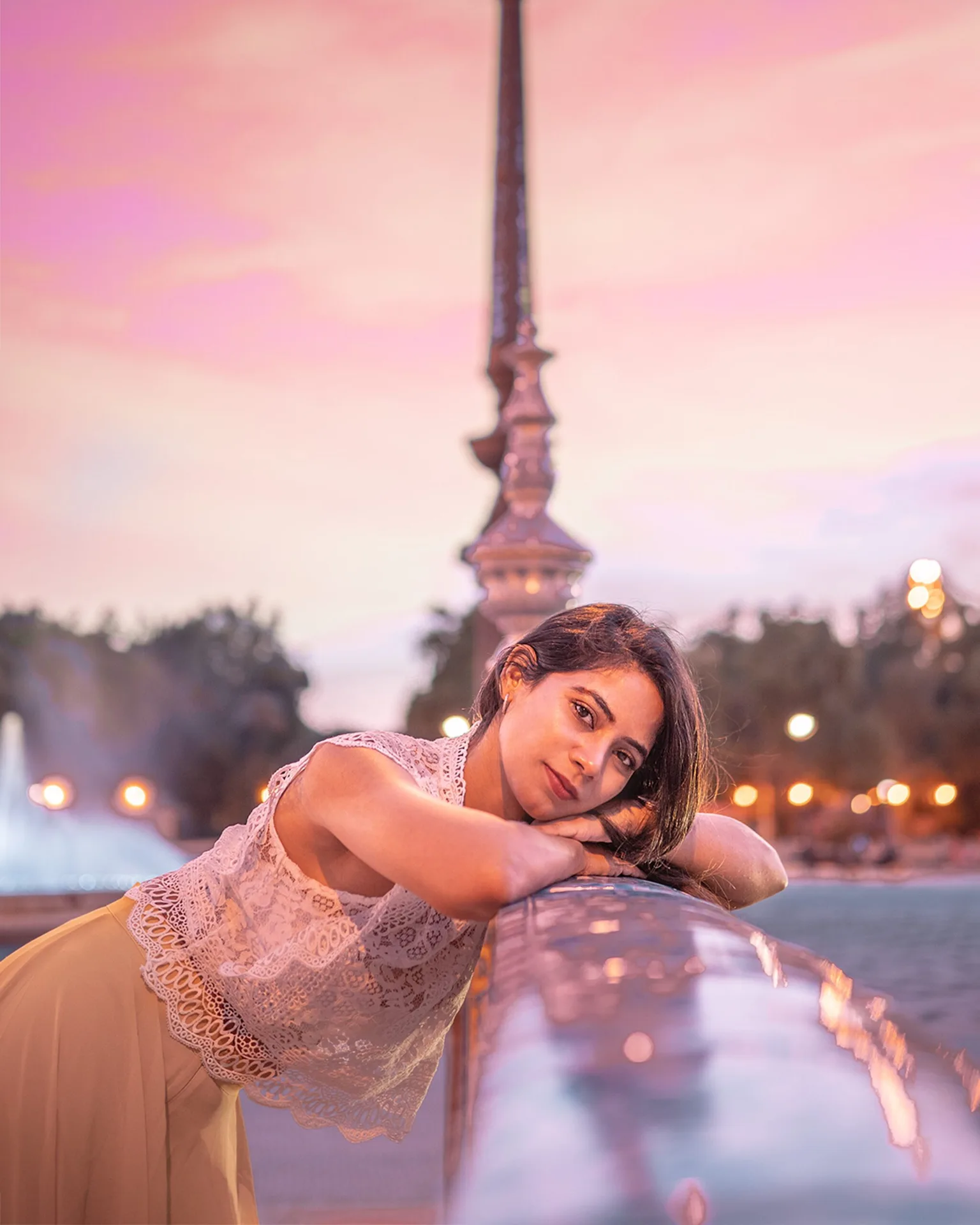Young woman in a lace top leaning on a bridge railing at dusk with pink skies and bokeh city lights behind her