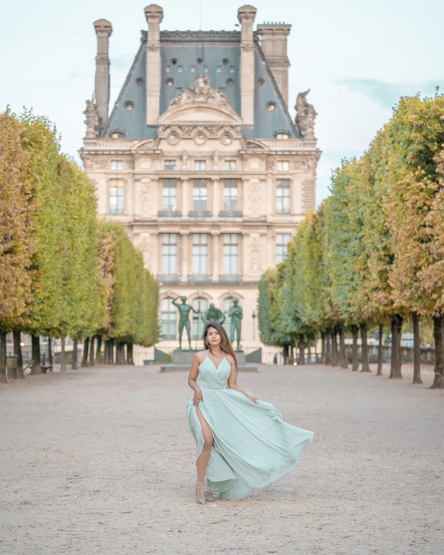 Woman in a flowing mint-green dress posing on a tree-lined path with a grand historic building in the background