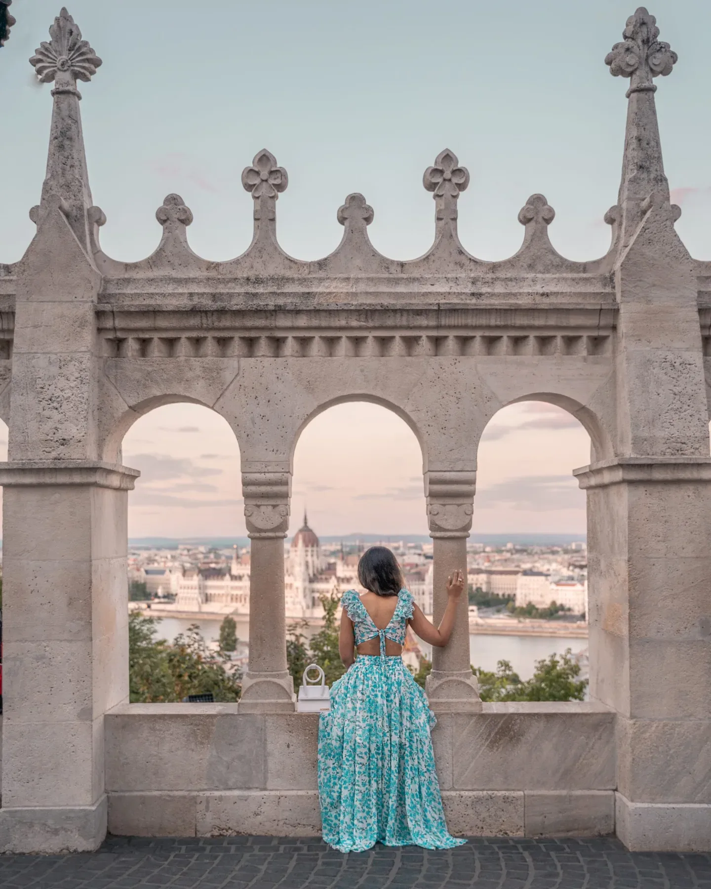 Woman in a turquoise floral dress standing at Fisherman's Bastion arches, overlooking the Budapest skyline and Parliament across the Danube.