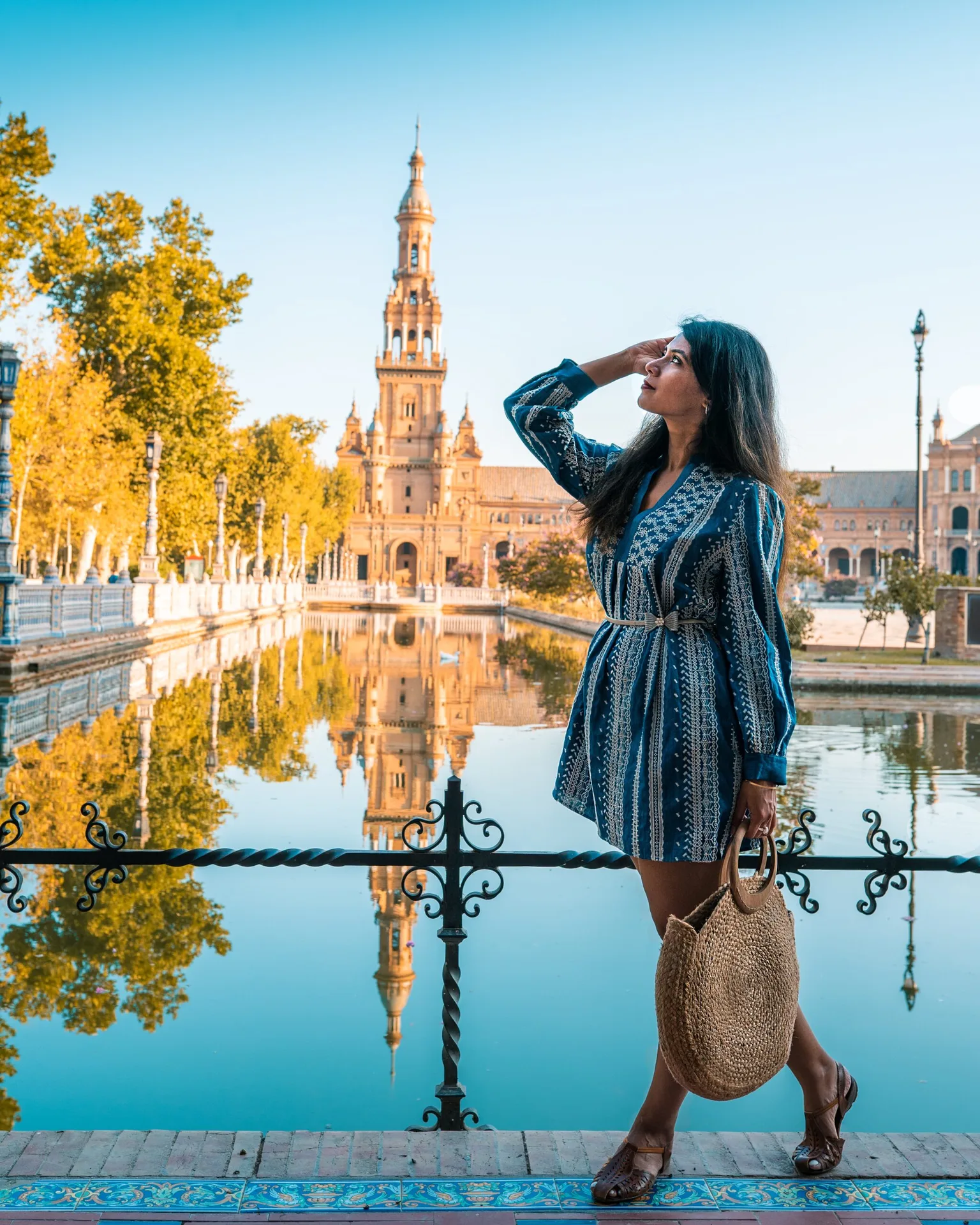 Woman in a blue patterned dress carrying a woven tote, standing by a reflective canal with ornate railing and a tall historic tower in the background.
