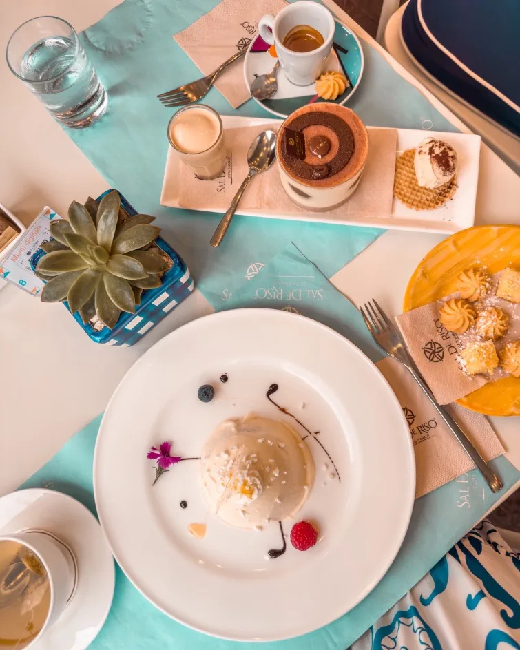 Plated panna cotta with berries and edible flower surrounded by espresso, petit desserts and a succulent on a turquoise tablecloth.