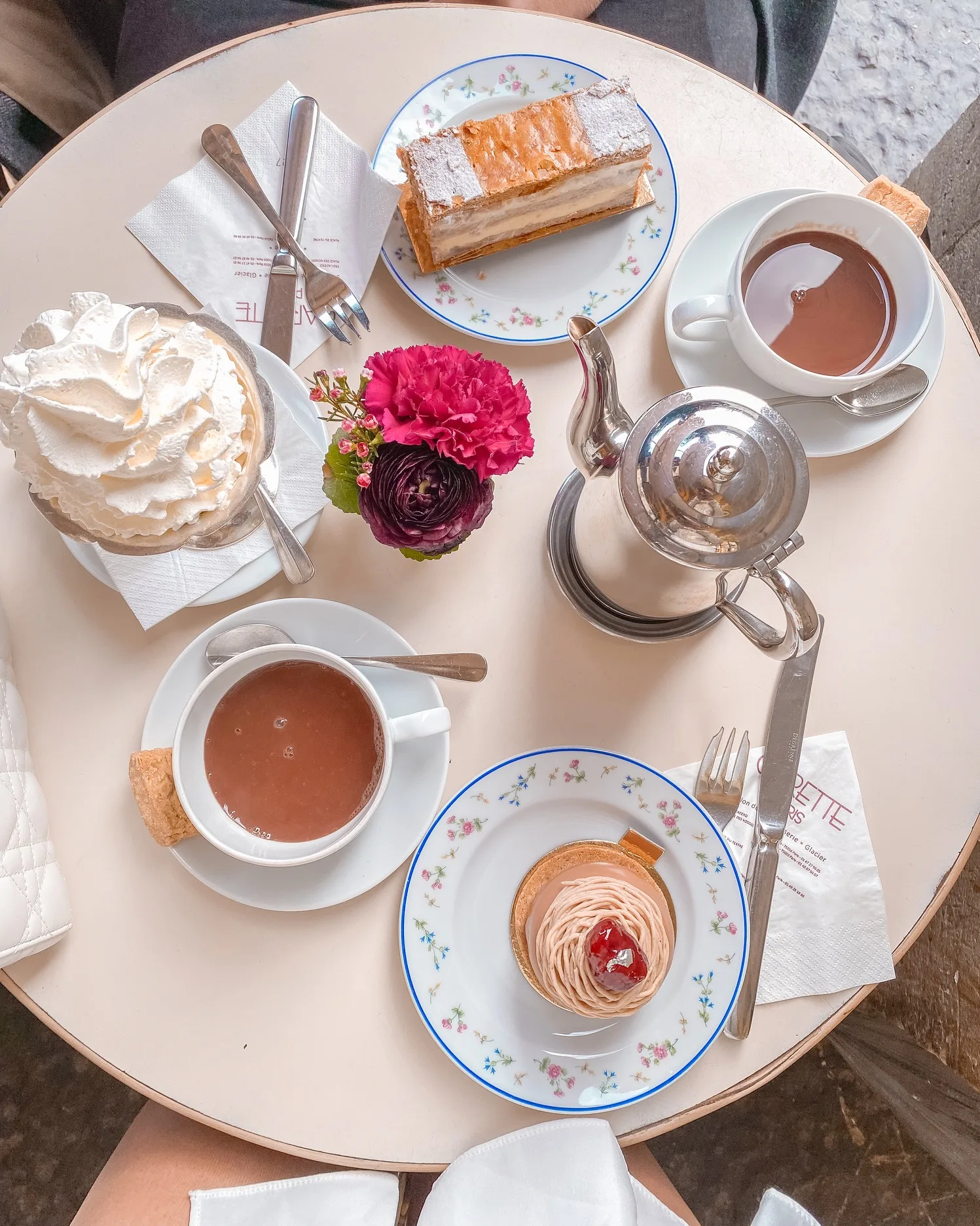Round café table set with two cups of hot chocolate, pastries on floral plates, a silver teapot and a small vase of pink flowers.
