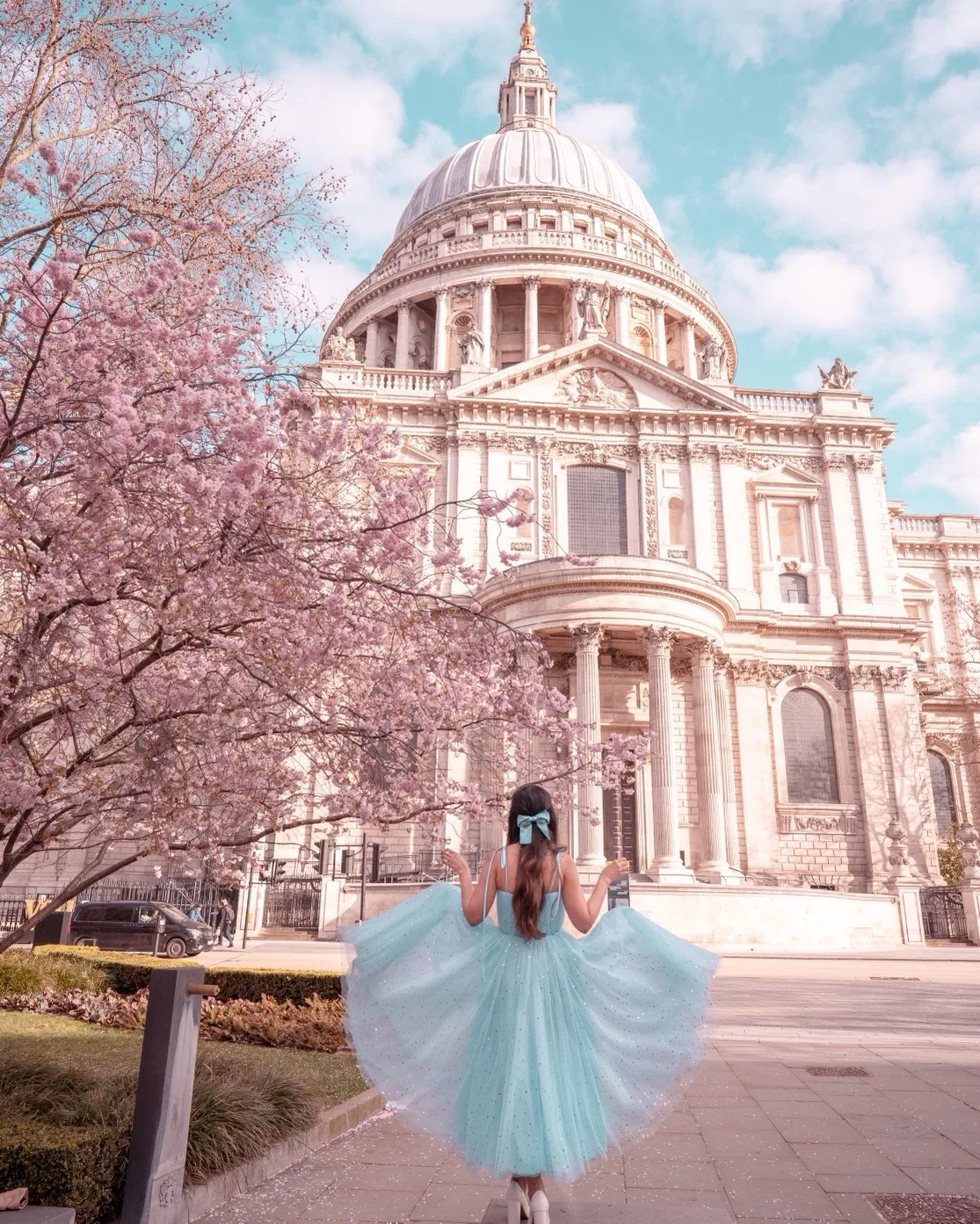 Woman in a pastel blue tulle dress and matching bow holding out her skirt beneath blossoming cherry trees in front of a grand domed cathedral.