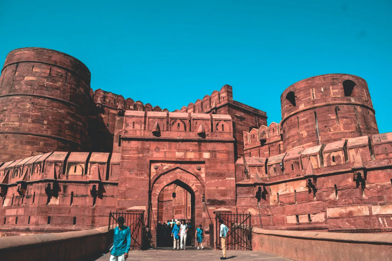 Massive red sandstone fort entrance with rounded towers and tourists passing through the arched gateway under a clear blue sky.