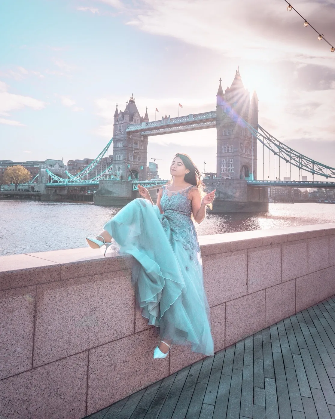 Woman in a flowing teal gown posing on a riverside wall with Tower Bridge and soft sunset light behind her.