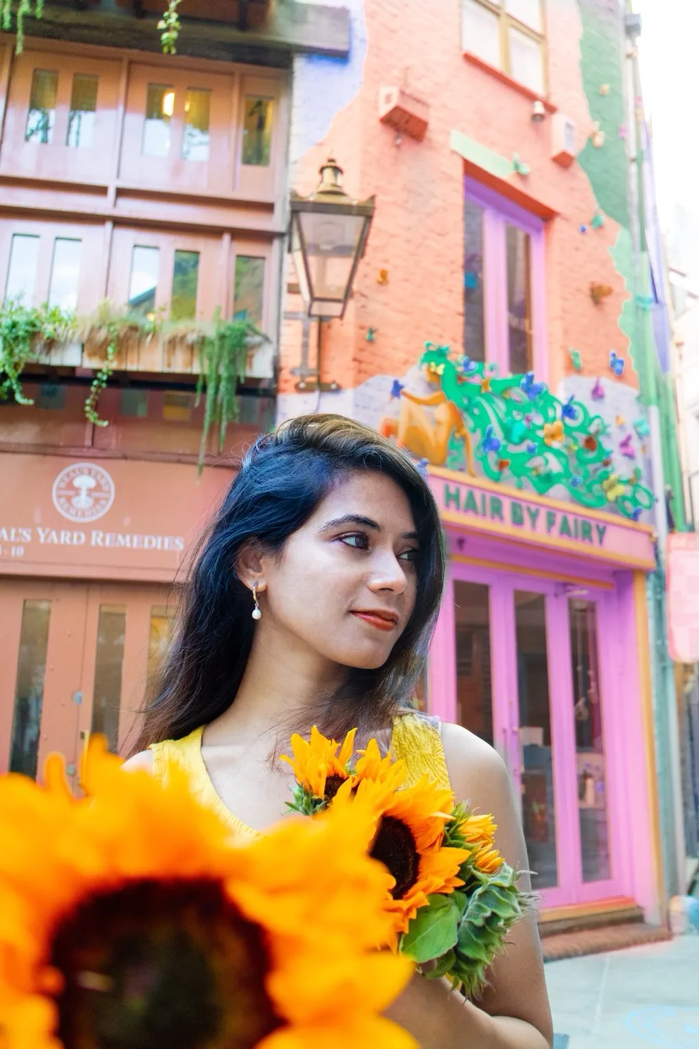 Young woman holding sunflowers in front of colourful shopfronts and a pink "Hair by Fairy" facade.