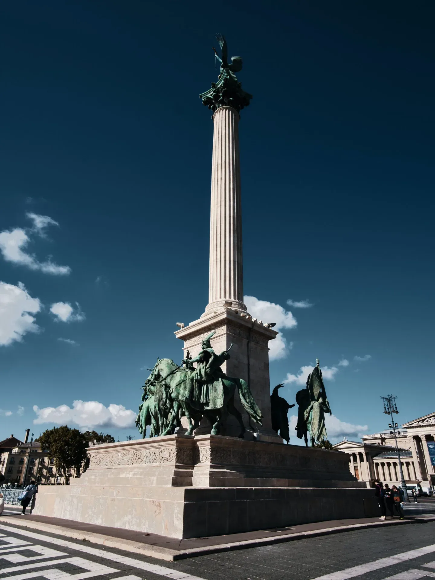 Tall stone column topped by a winged statue rising above bronze equestrian figures on a grand city square under a deep blue sky.