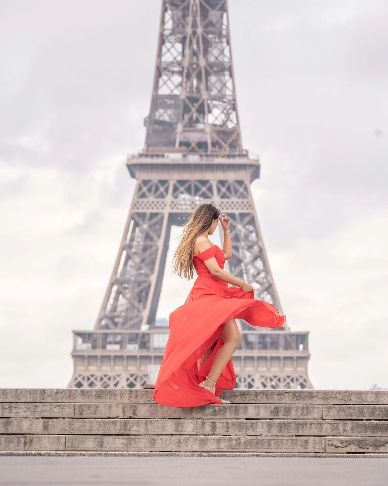 Woman in a flowing red dress posing on stone steps with the Eiffel Tower rising behind her on a cloudy day.