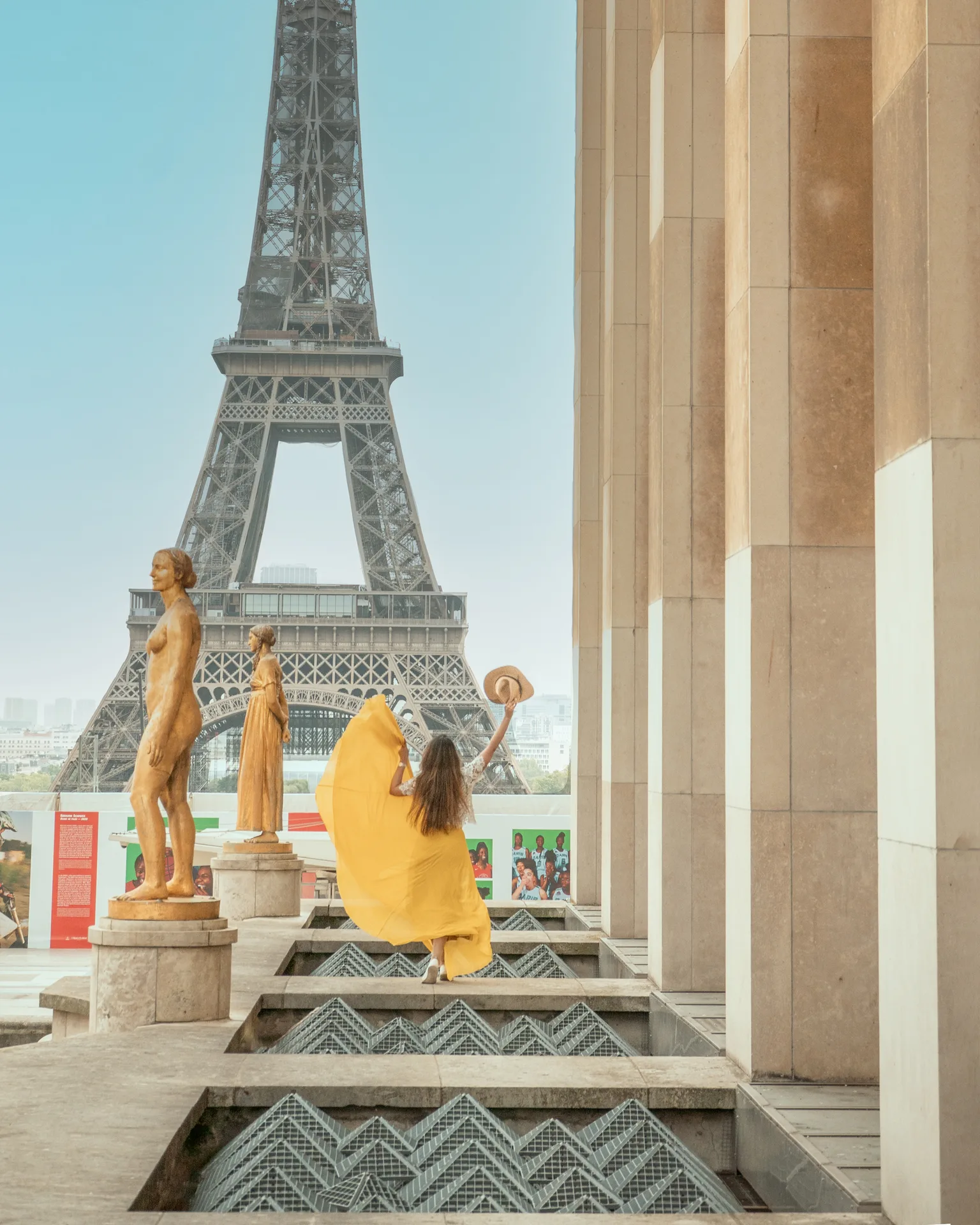 Woman in a flowing yellow dress and sunhat raised in front of the Eiffel Tower, framed by classical statues and stone columns.