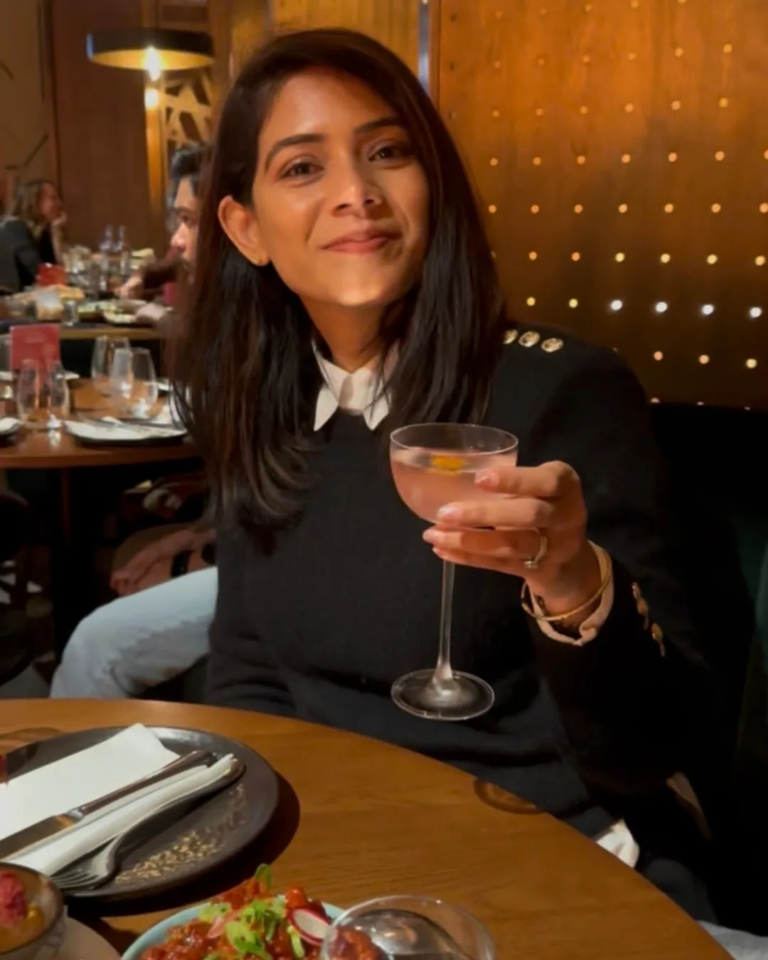 Woman smiling and raising a cocktail glass while seated at a warmly lit restaurant table with plates and shared dishes.