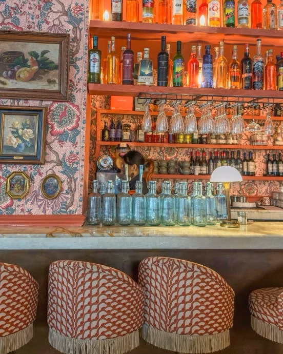 A stylish bar with patterned wallpaper, shelves of bottles and glassware, and fringed stools in the foreground