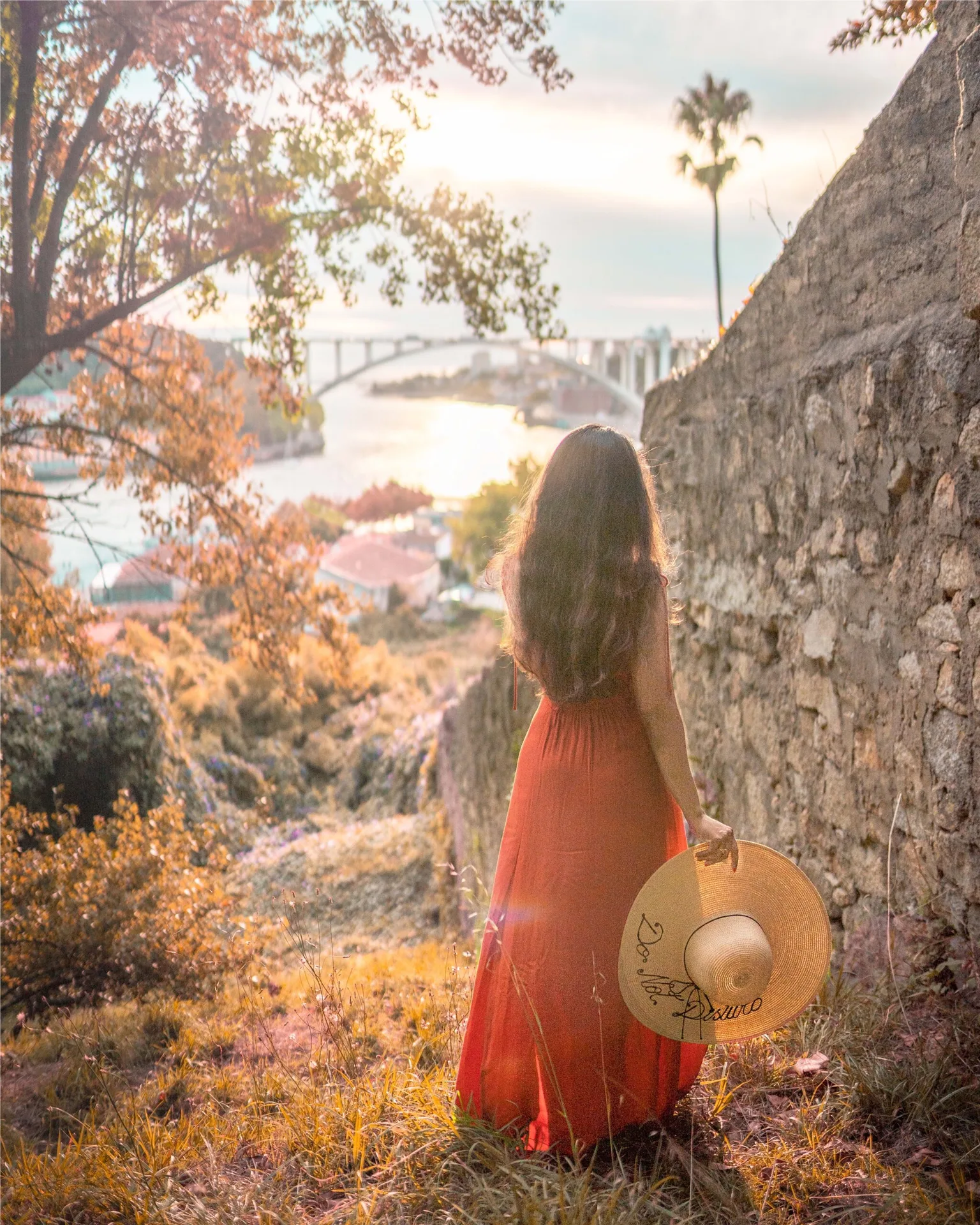 Woman in a flowing red dress holding a sun hat and gazing at a river and arched bridge at sunset.