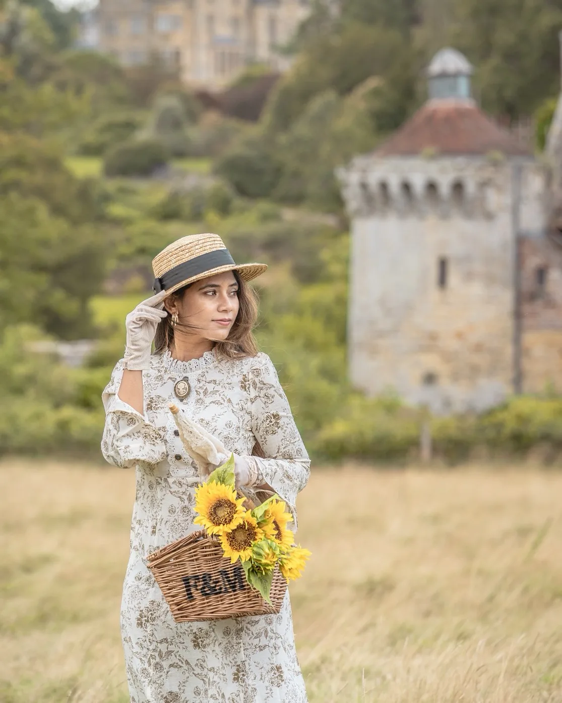 Woman in a vintage floral dress and straw boater hat holding a basket of sunflowers in a grassy meadow with a stone tower in the background.