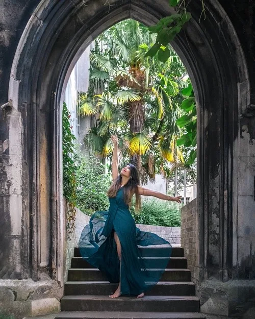 Woman in flowing teal dress poses barefoot on stone steps under a gothic arch with tropical palms beyond.