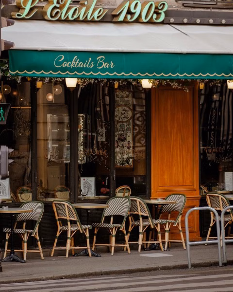 Street-side café and cocktail bar with a green awning and outdoor wicker tables in front of a Parisian-style façade