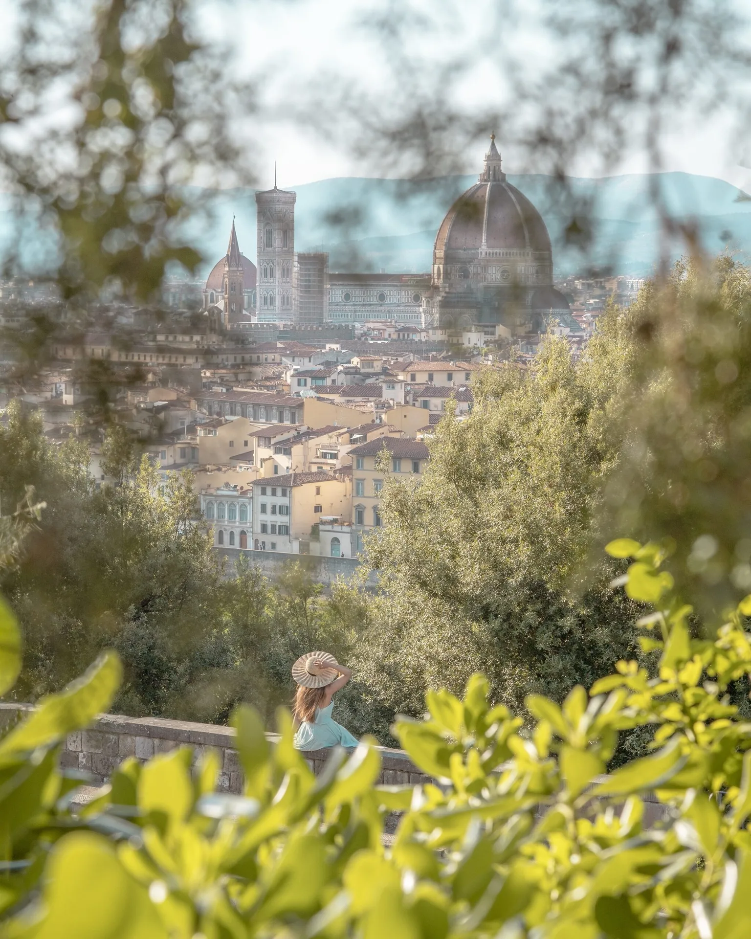 Woman in a sunhat sitting on a ledge overlooking Florence with the Duomo and city rooftops framed by leafy trees.