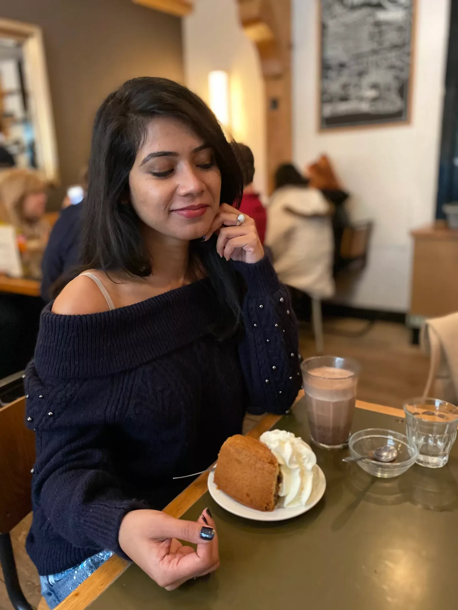 Woman in a cosy café smiling at her cake and whipped cream beside a glass of hot chocolate.