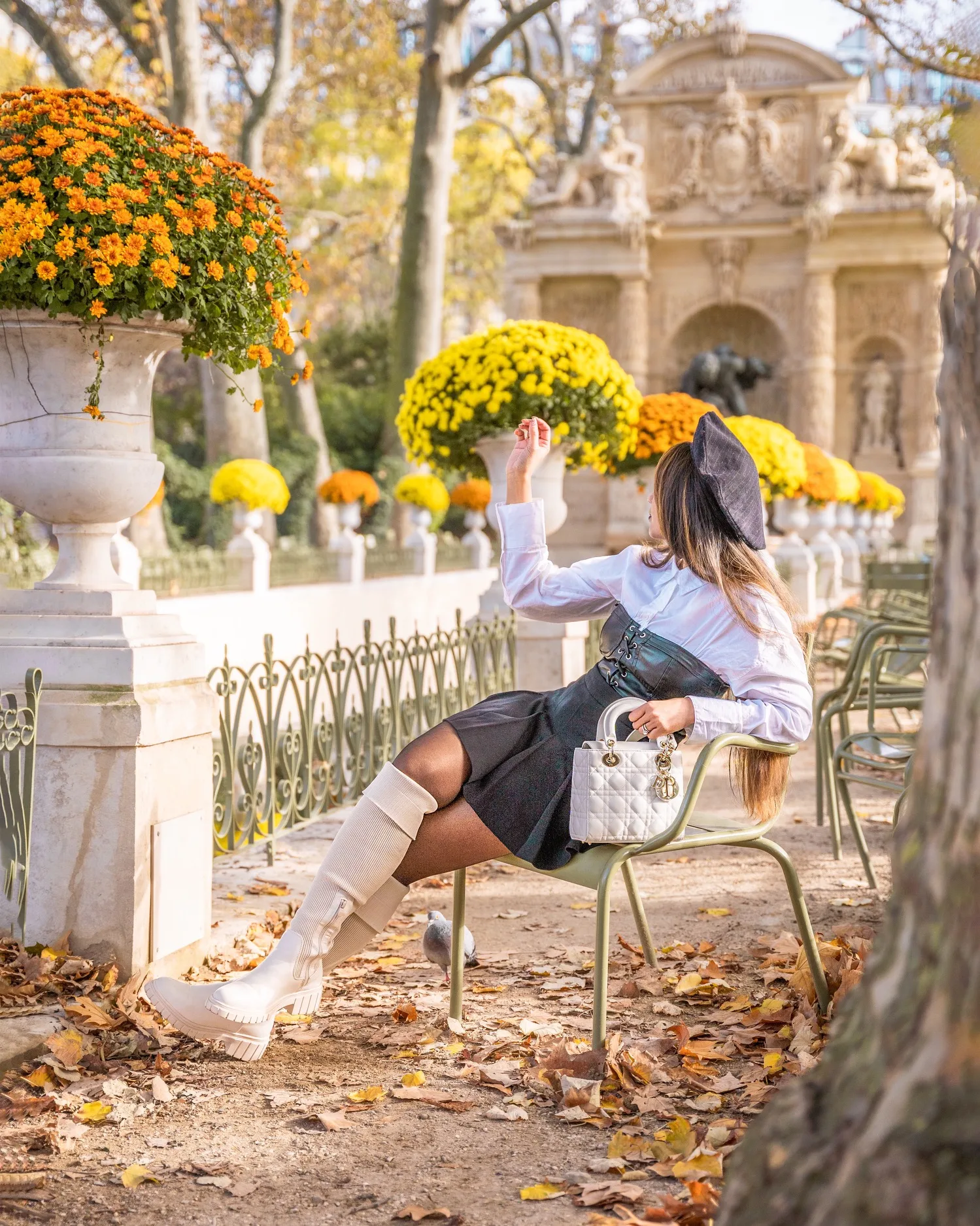 Woman in a beret and knee-high boots lounging on a green park chair beside urns of yellow and orange chrysanthemums in an ornate autumn garden