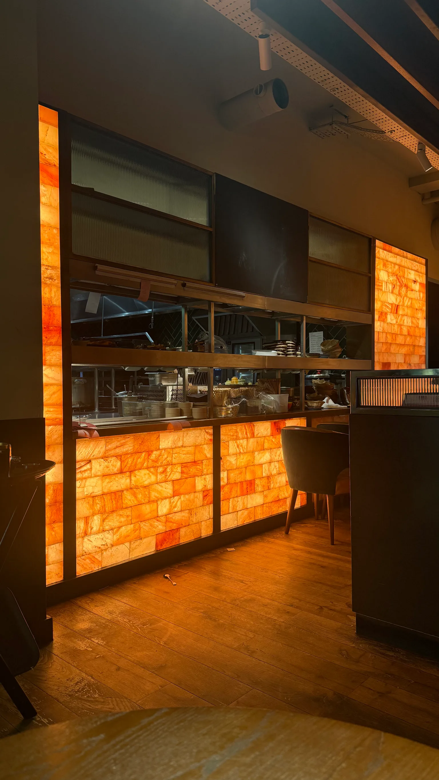 Warm-lit restaurant pass with glowing orange stone panels, a view into the busy kitchen and wooden floor.