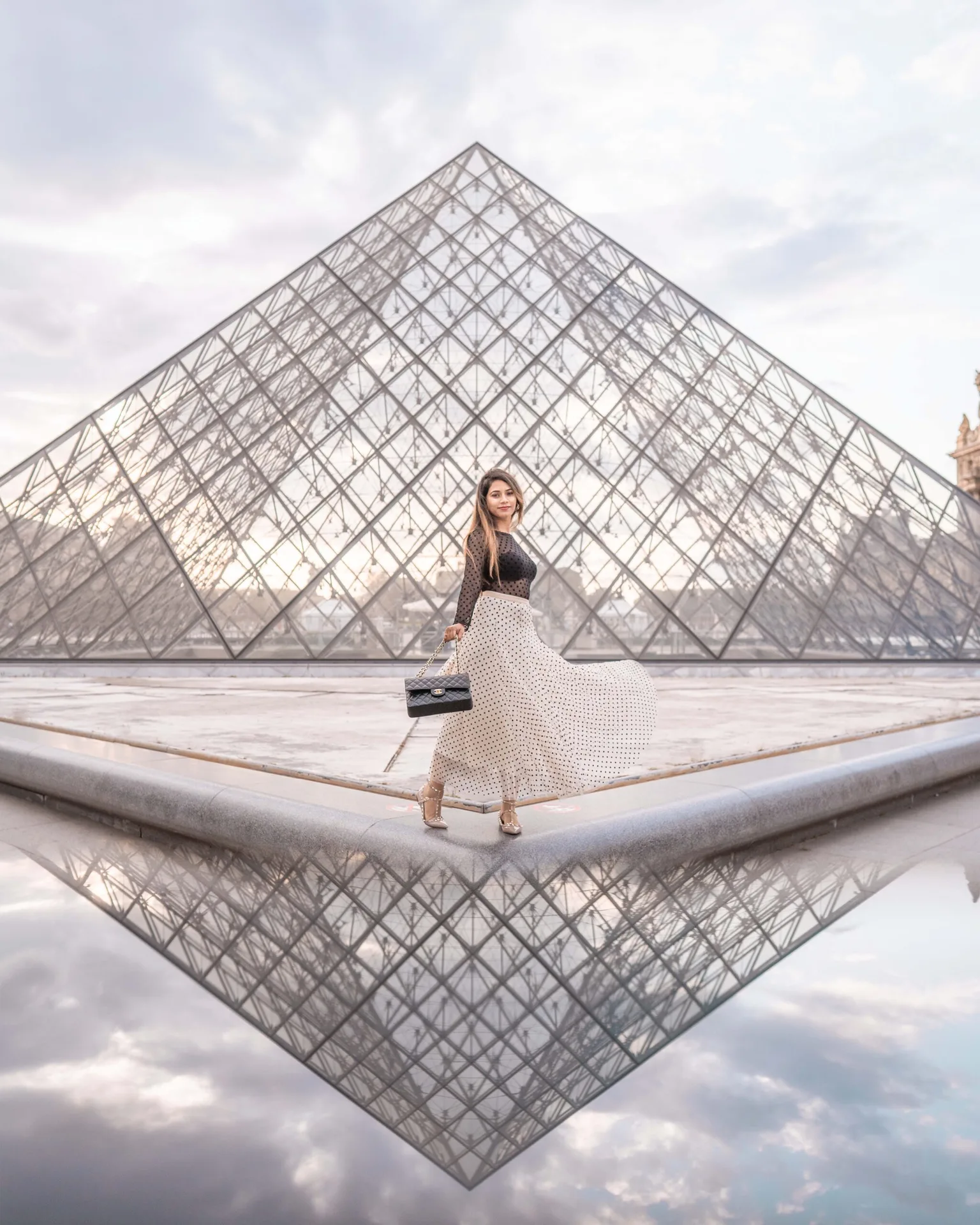 Woman in a flowing polka-dot skirt posing with a handbag in front of the Louvre glass pyramid, its reflection visible in the foreground