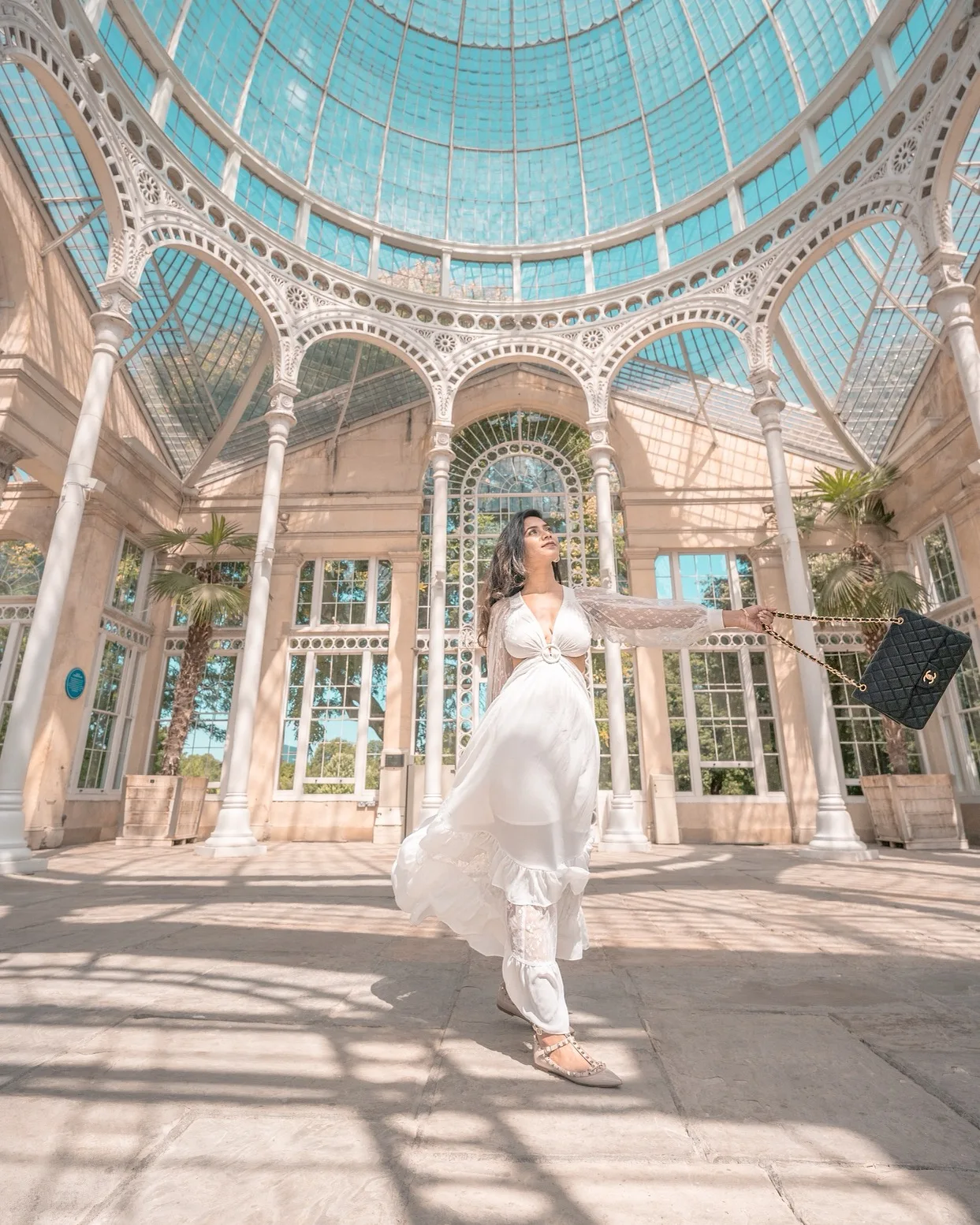 Woman in a flowing white dress twirling beneath a glass-domed Victorian conservatory with ornate iron arches.