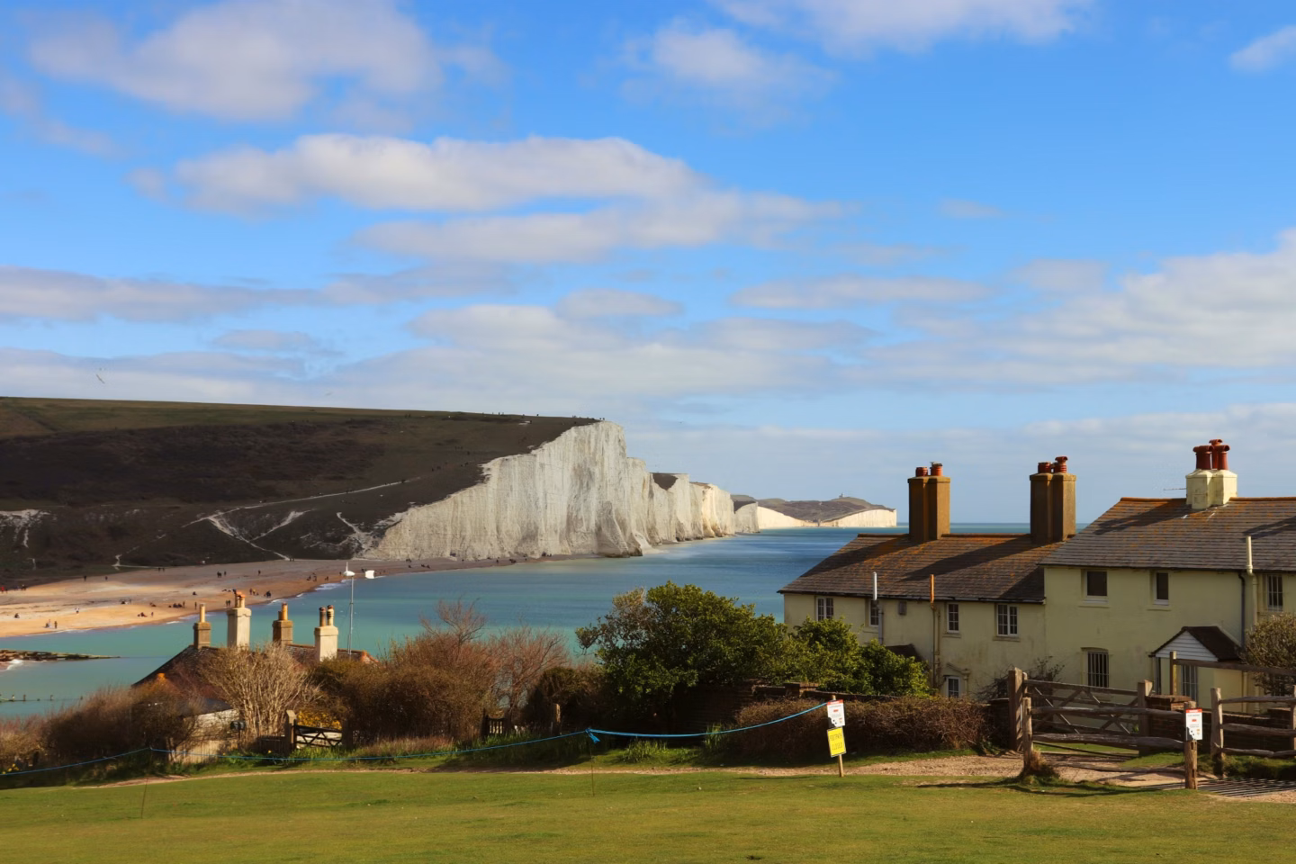 White chalk cliffs beside a turquoise bay, viewed past seaside houses under a blue sky