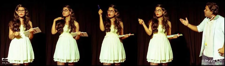 Woman in a white dress and glasses making animated gestures on a dark stage while a man in casual clothes stands to the side.