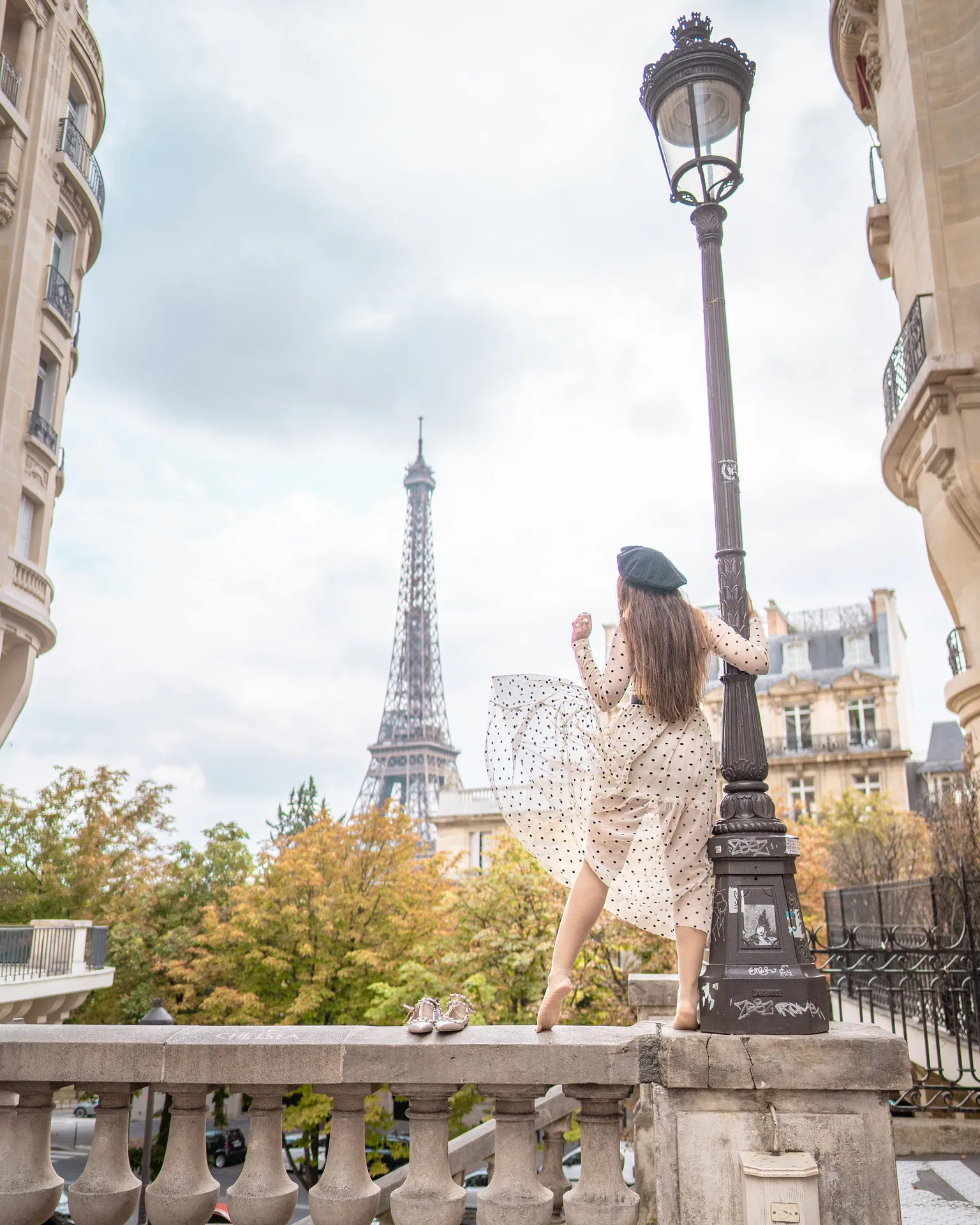 Woman in a polka-dot dress and beret posing on a Paris balcony with the Eiffel Tower in the background.
