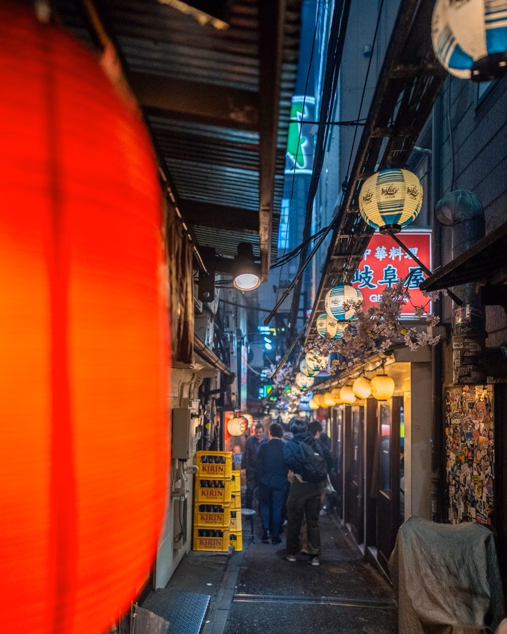 Narrow alleyway lit by lanterns and neon signs, with people walking past bars and restaurants at night