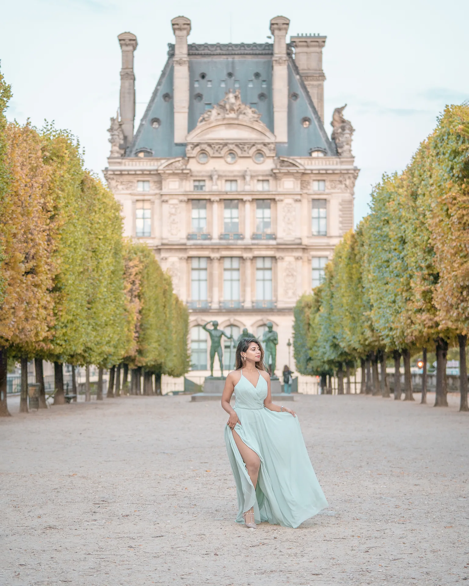 Woman in a flowing pale green dress walking down a tree-lined path with a grand historic building and statues in the background