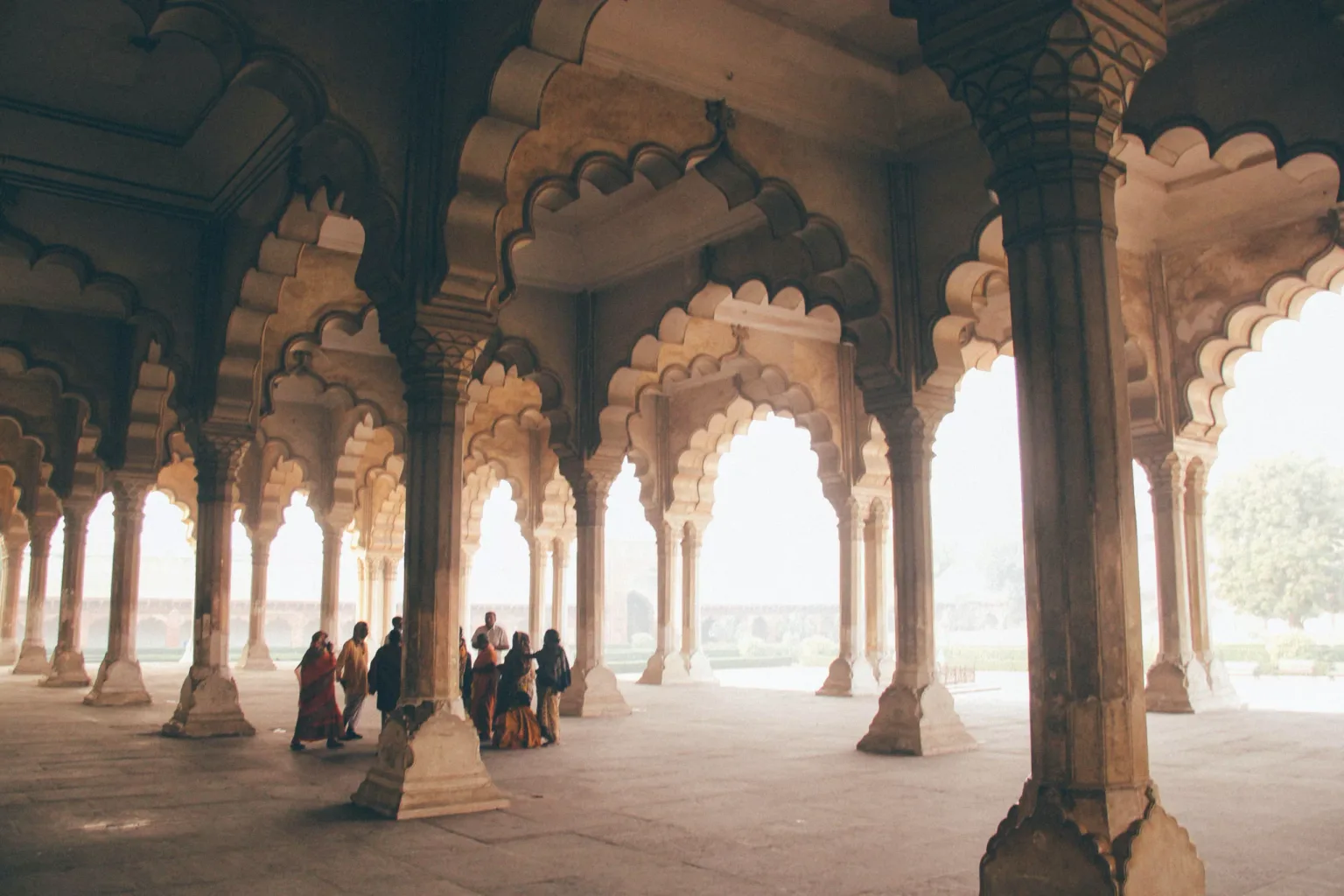 Sunlit Mughal-style hall with scalloped arches and a small group of visitors gathered near its central columns.