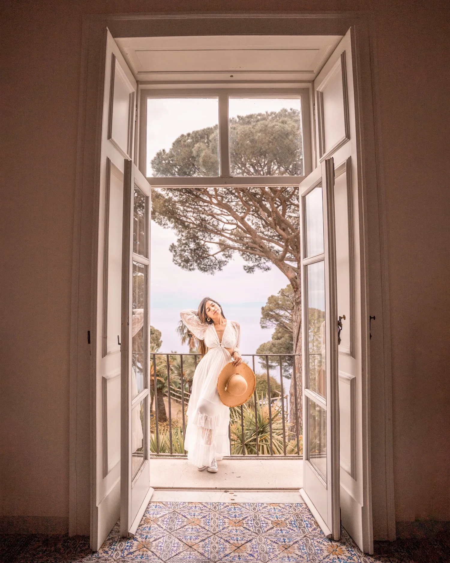 Woman in a flowing white dress holding a wide-brimmed hat, standing on a balcony framed by open French doors with pine trees and sea beyond