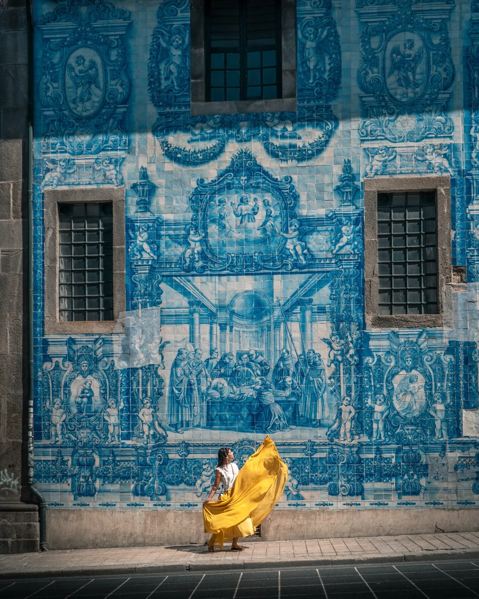 Woman in a flowing yellow skirt posing against a large blue-and-white azulejo-tiled mural on a city street.
